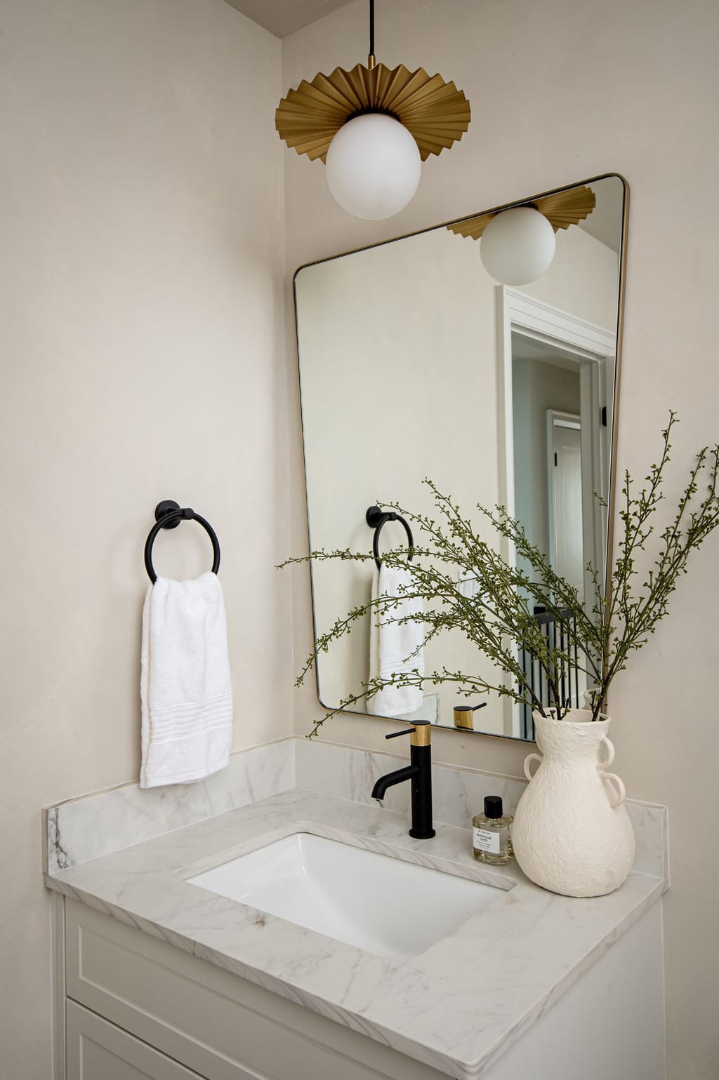 A small bathroom vanity with a marble top, black faucet, brass-framed mirror and a pleated brass globe light