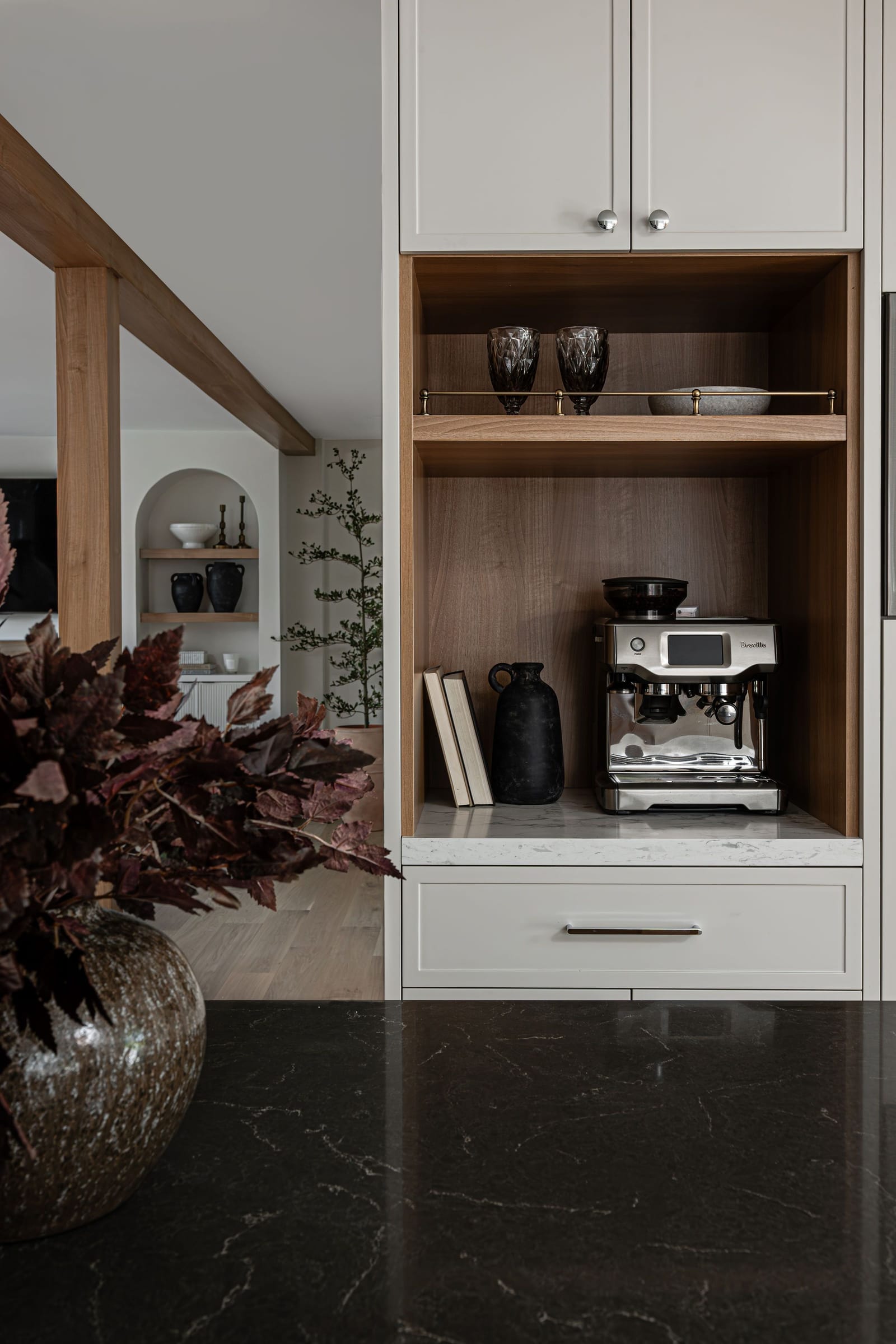 Close view of the oak-lined coffee niche with an espresso machine, cups on a shelf with a thin brass rail and a lit underside