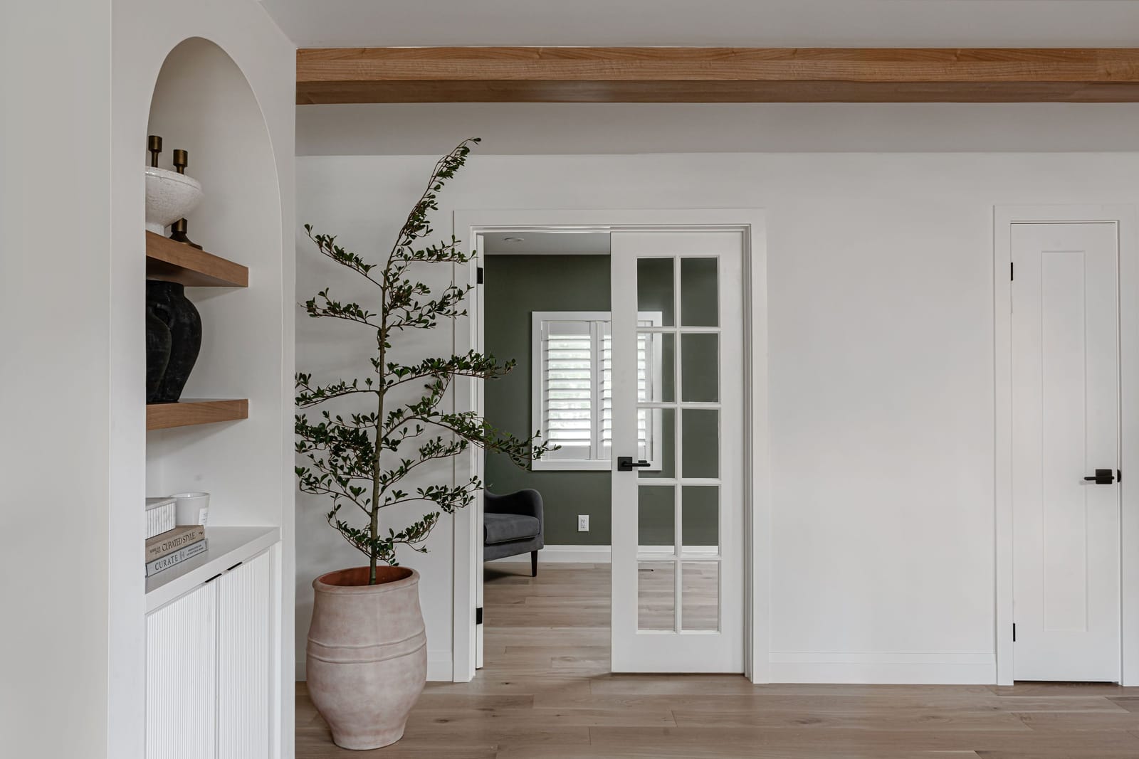 The living room with an arched niche of oak shelves, a terracotta pot with an olive tree and French doors through to the green office