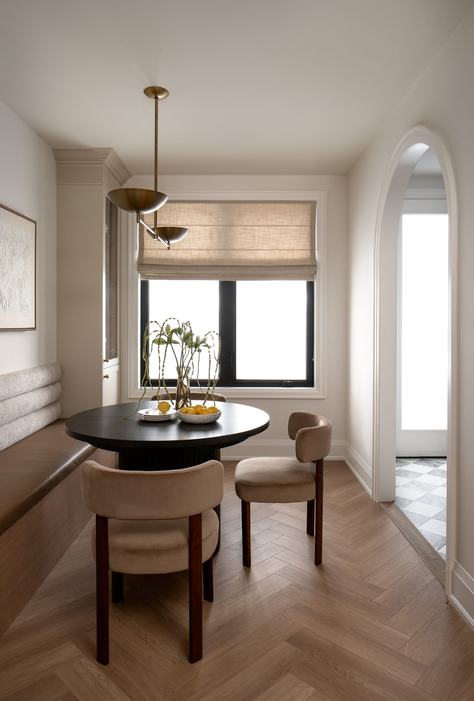 The dining nook seen from the side, with a linen blind over the window and an arched opening through to a checkerboard-floored mudroom
