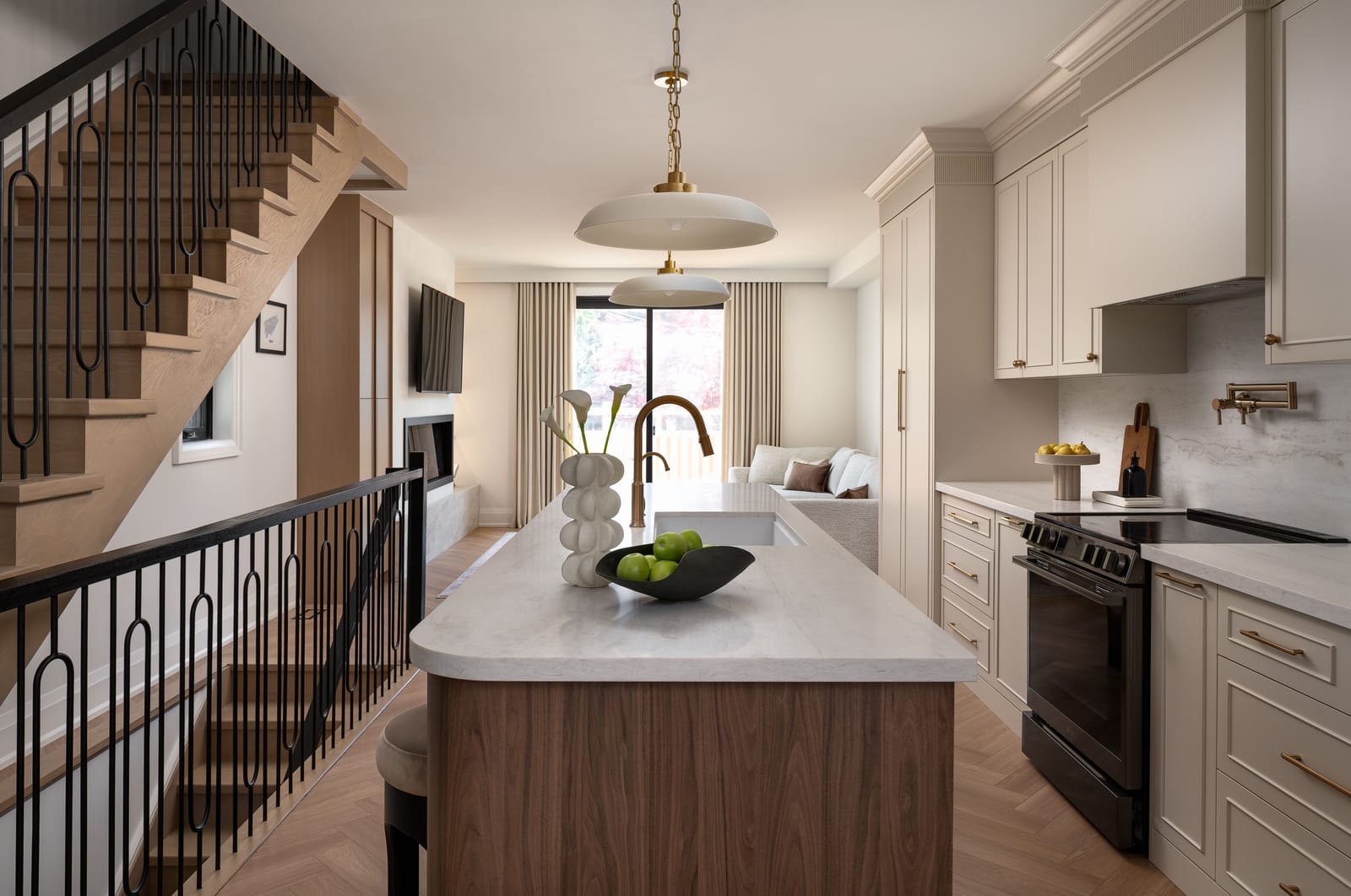 A narrow kitchen with a walnut island, cream shaker cabinets and a herringbone floor, beside an open oak stair with looped black balusters