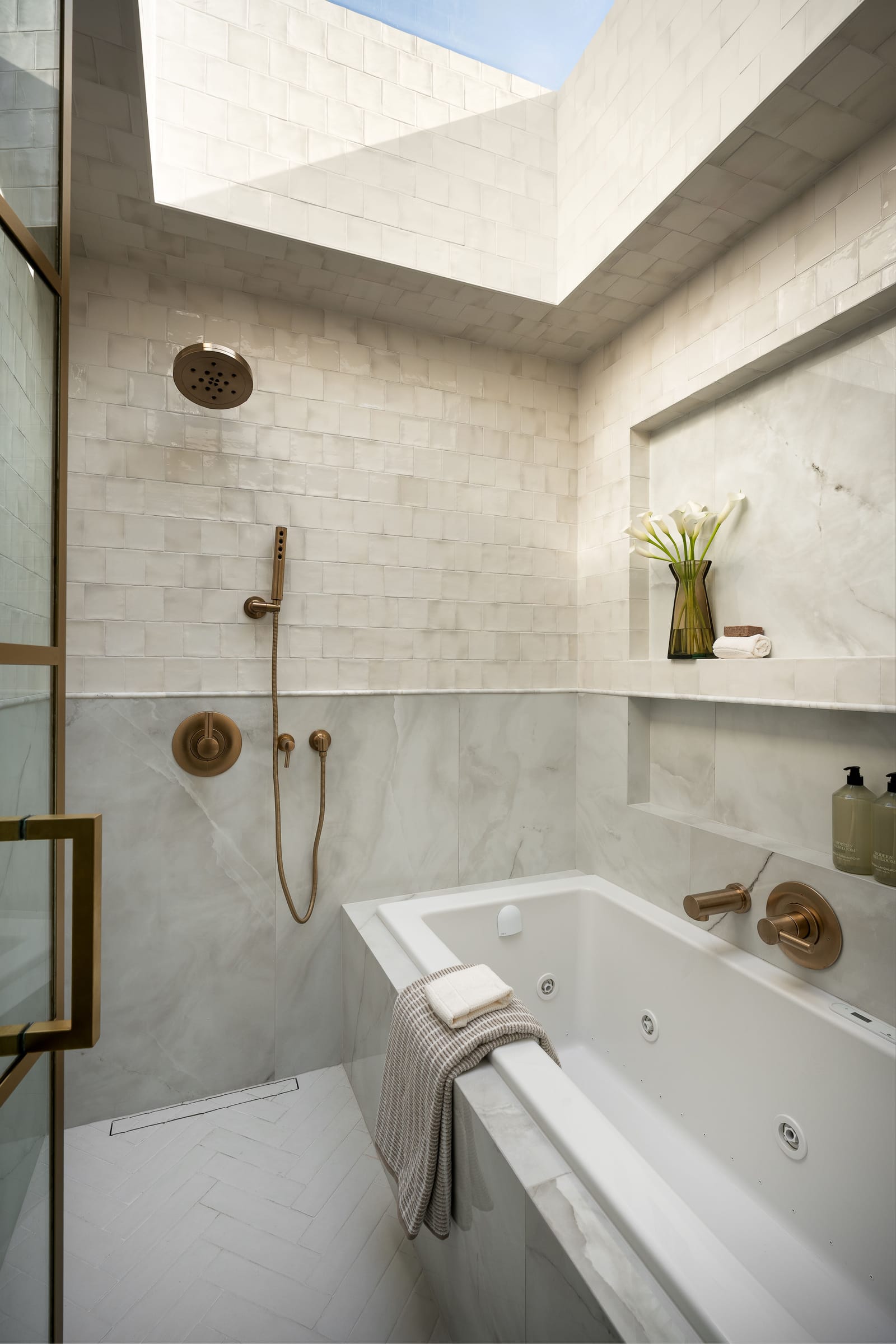 A wet room lit by a skylight, with zellige-style wall tile, marble-look slabs, a freestanding tub, a brass rain shower and a linear drain