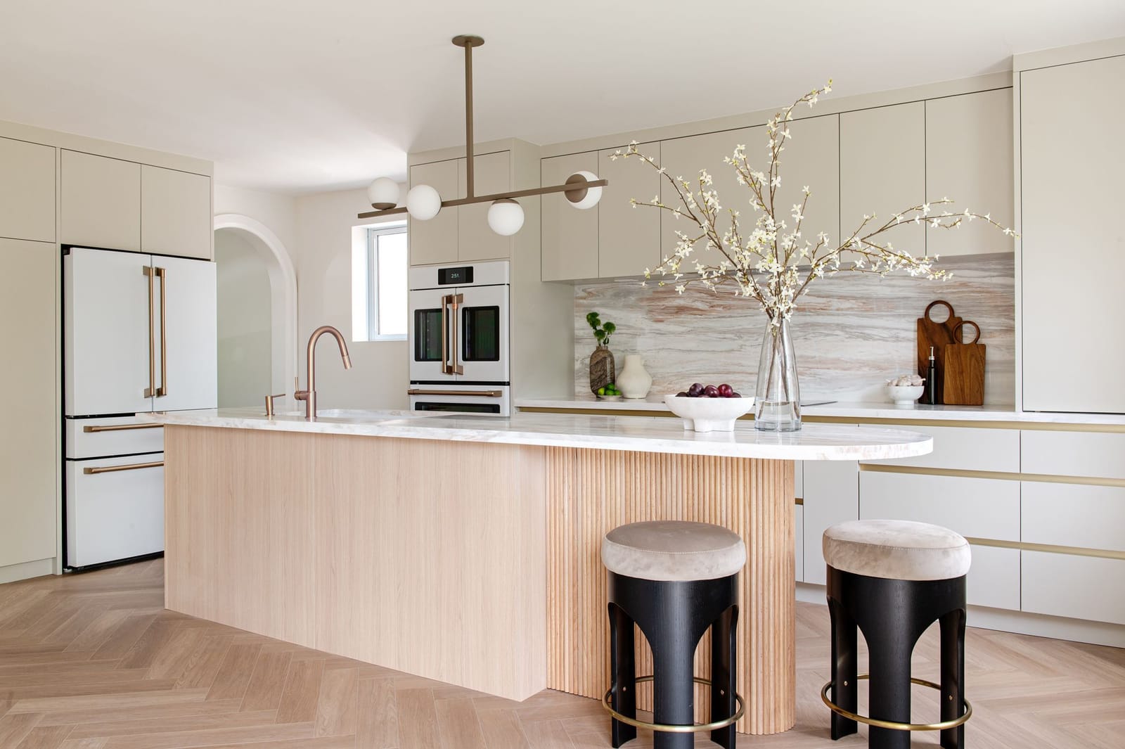 A cream kitchen with a fluted pale oak island under a pink-veined stone top, black and brass stools, a herringbone floor and white wall ovens