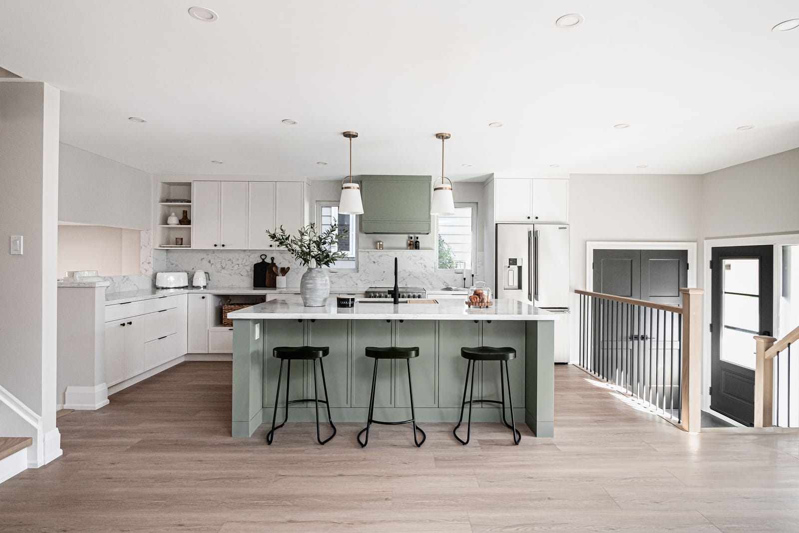 An open kitchen with a sage-green island and hood, white shaker cabinets, black stools and an oak and black-spindle stair rail to the right